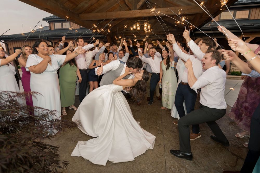 Bride and groom share a romantic dip kiss surrounded by cheering guests holding sparklers at outdoor wedding reception.