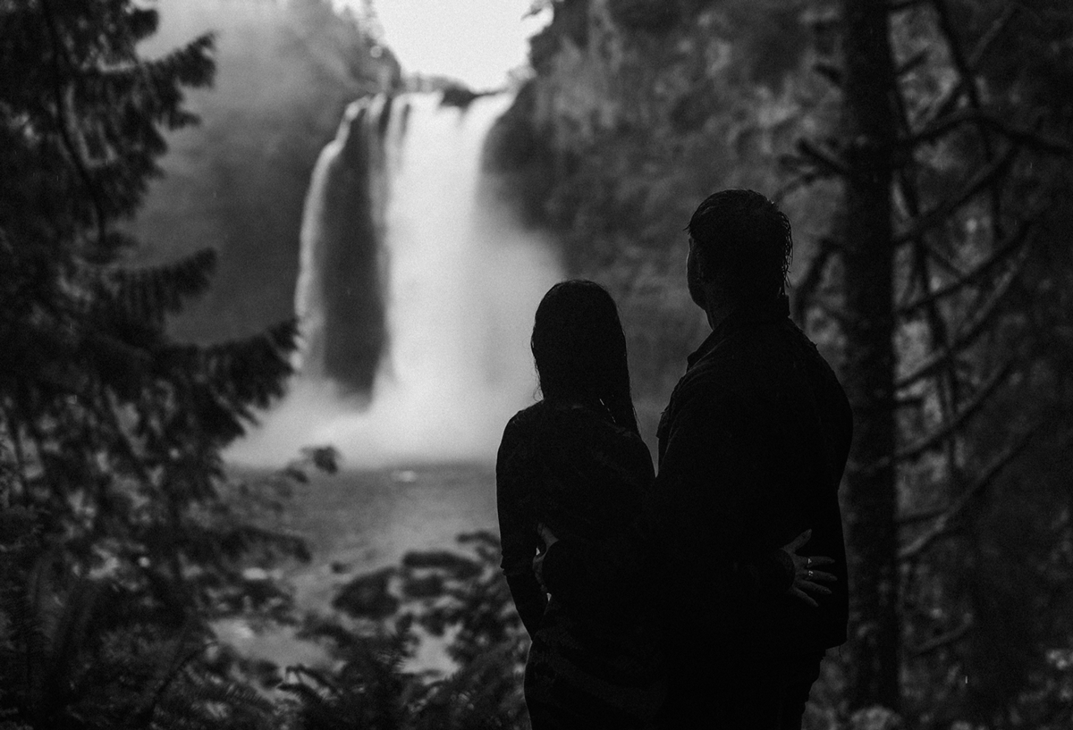 Silhouette of a couple standing together admiring a majestic waterfall in a misty forest, black and white photo.