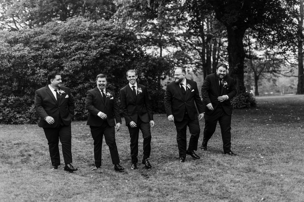 Black and white photo of a groom and five groomsmen in suits walking together outdoors on a grassy lawn.