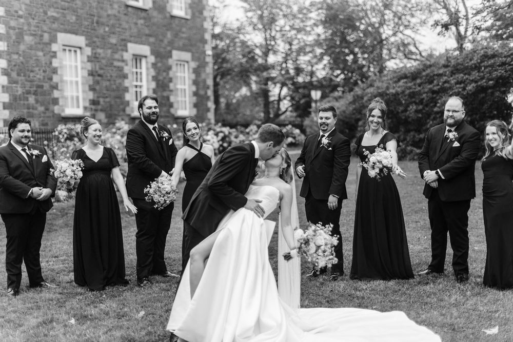 Black and white photo of bride and groom kissing surrounded by wedding party on lawn outside stone building.