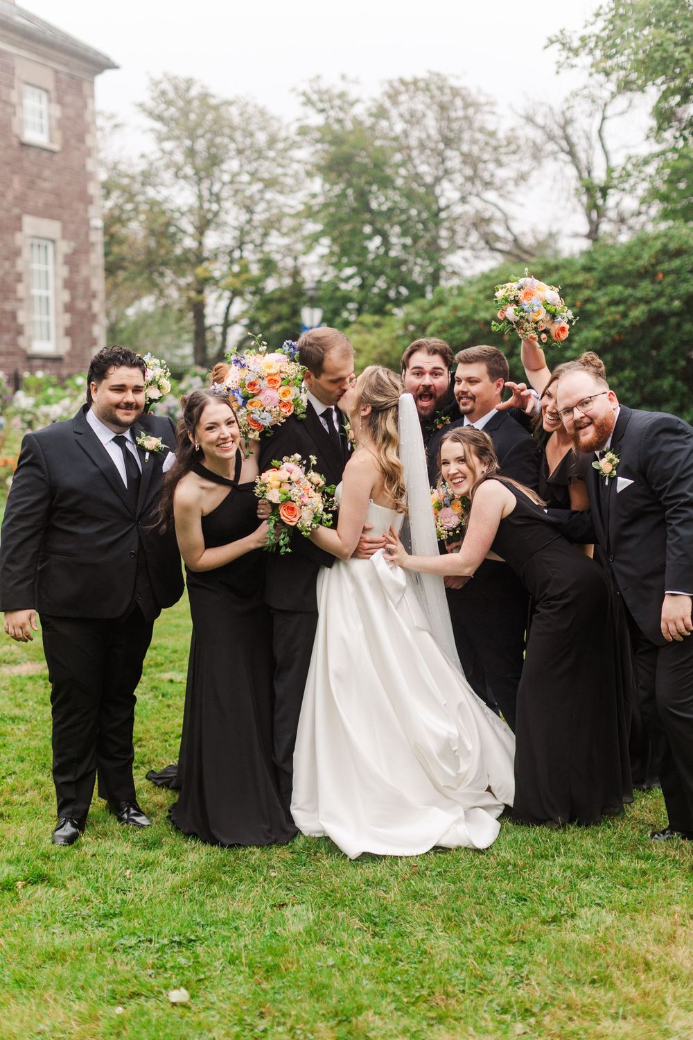 Wedding party in black attire surrounds smiling bride and groom on green lawn outside historic stone building.