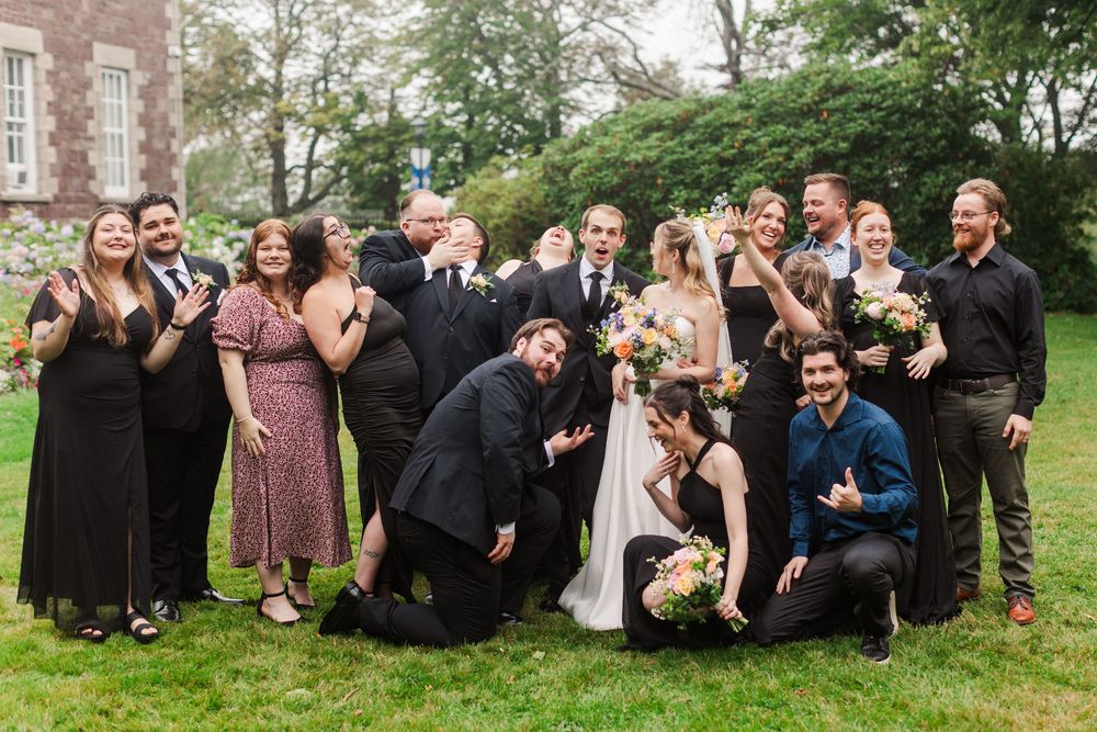 Wedding party posing playfully on green lawn, bride in white gown holding colorful bouquet surrounded by guests in formal attire.