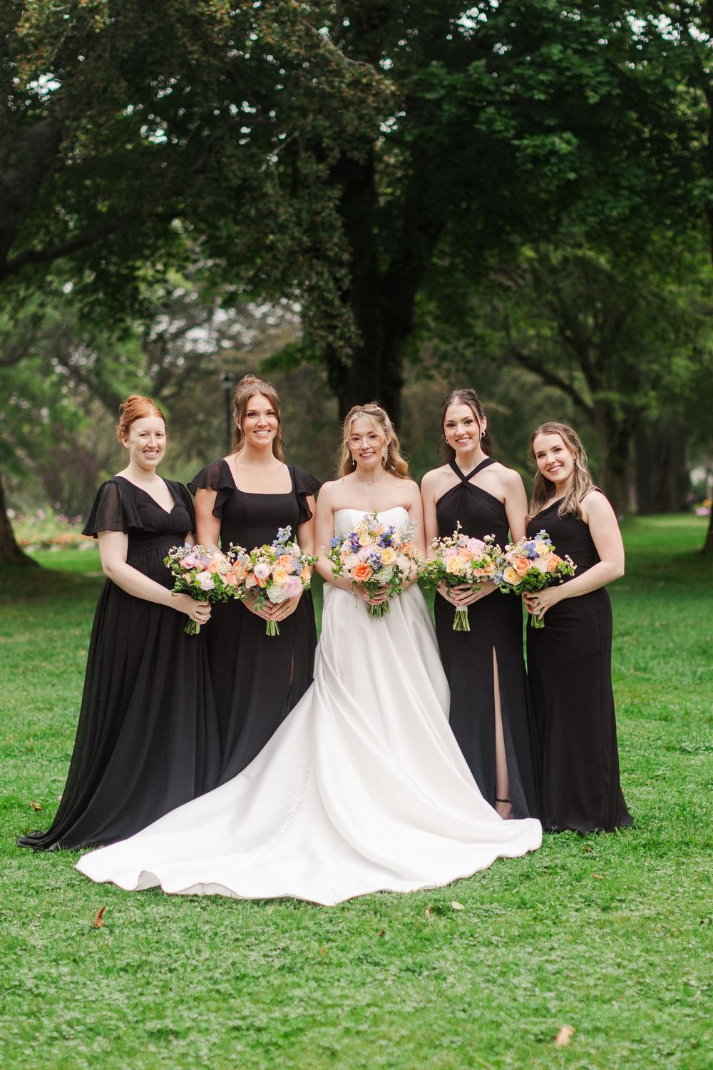 Bride in white gown poses with four bridesmaids in black dresses holding colorful bouquets on green lawn.