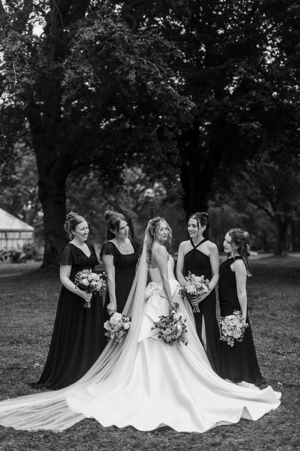Black and white photo of a bride in a white gown with four bridesmaids in dark dresses holding bouquets outdoors.