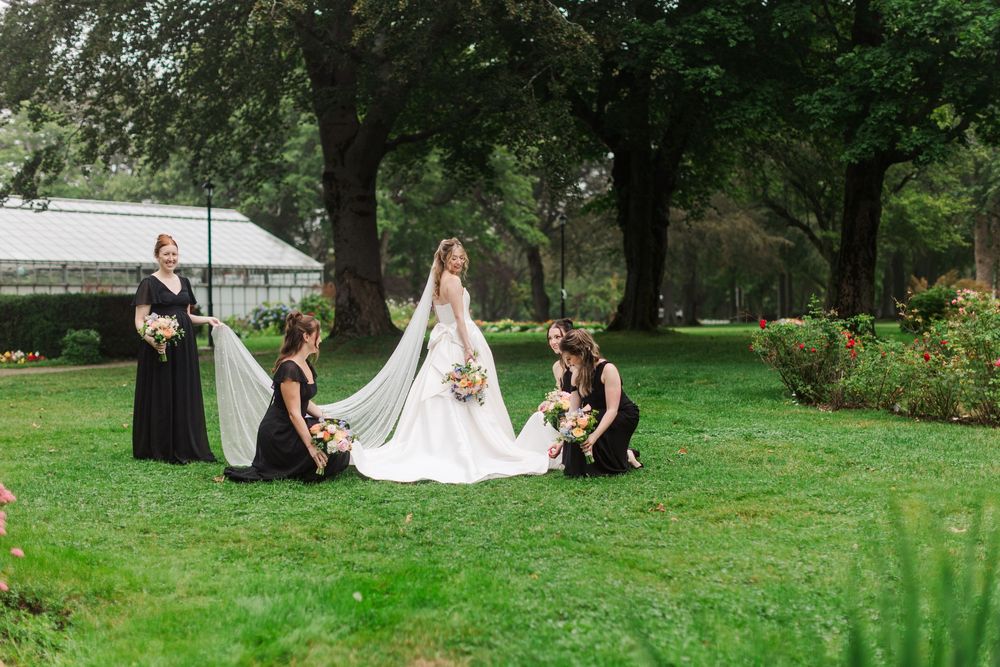 Bride in white gown with bridesmaids in black dresses arranging her train on lush green lawn near trees.