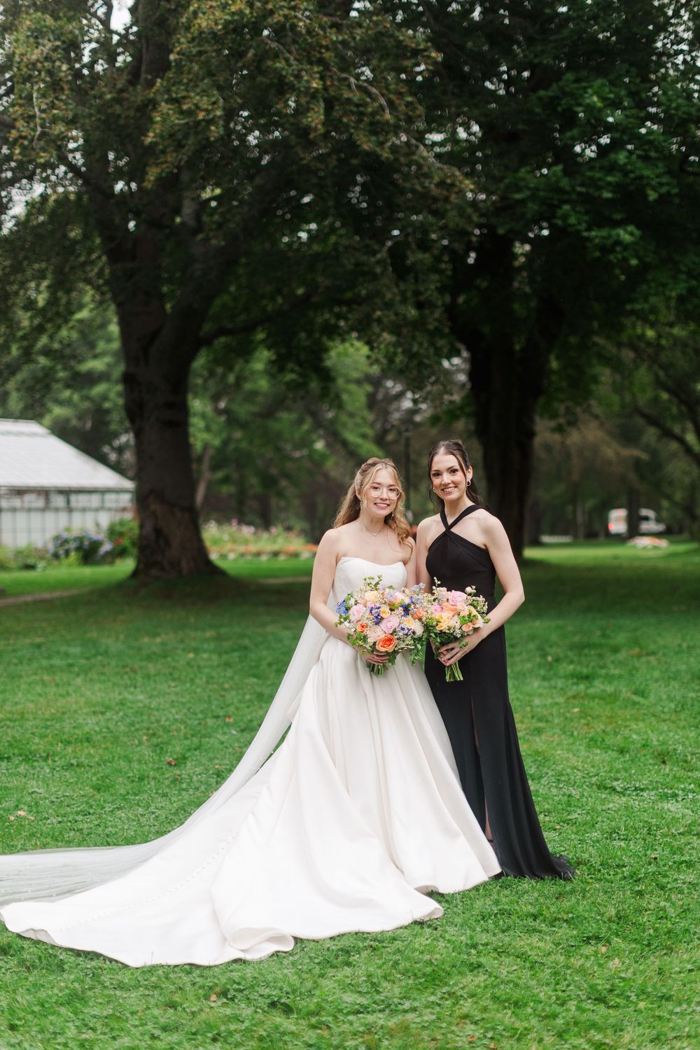 Bride in white gown and bridesmaid in black dress holding colorful bouquets, posing on green lawn with trees.