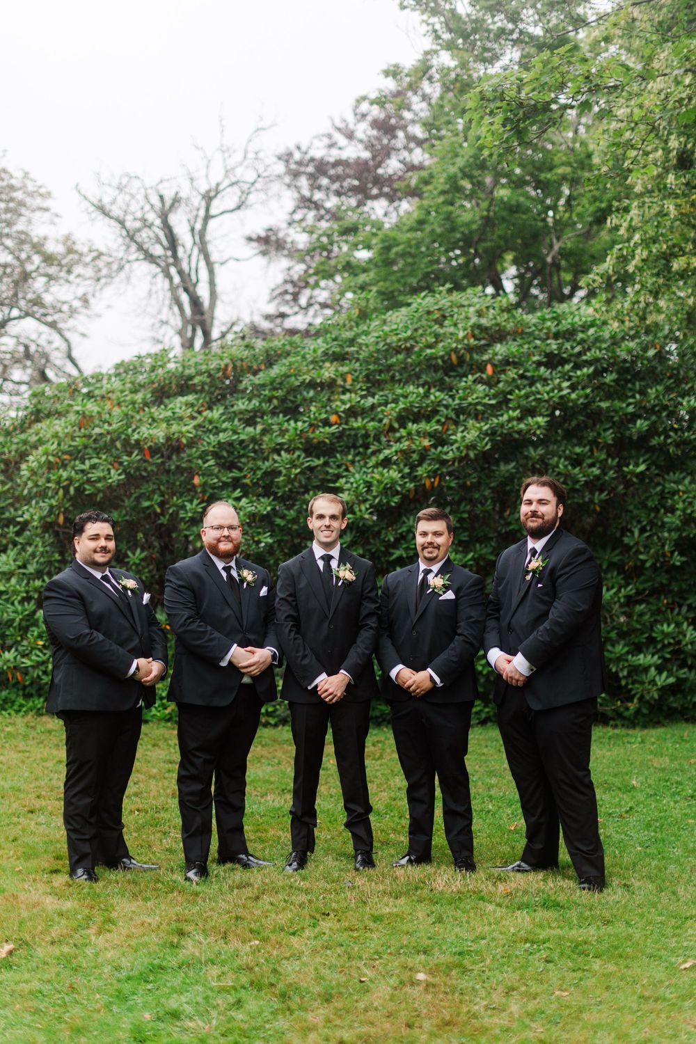 Five groomsmen in black suits with boutonnieres pose on green lawn before lush hedge backdrop at outdoor wedding.