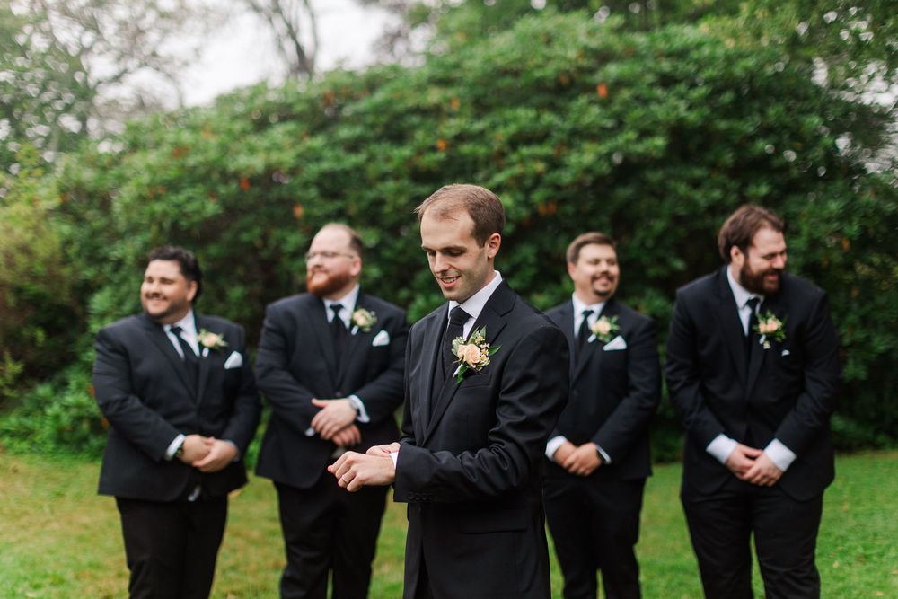 Groom adjusting cufflinks in black suit with groomsmen standing behind him outdoors at a wedding.