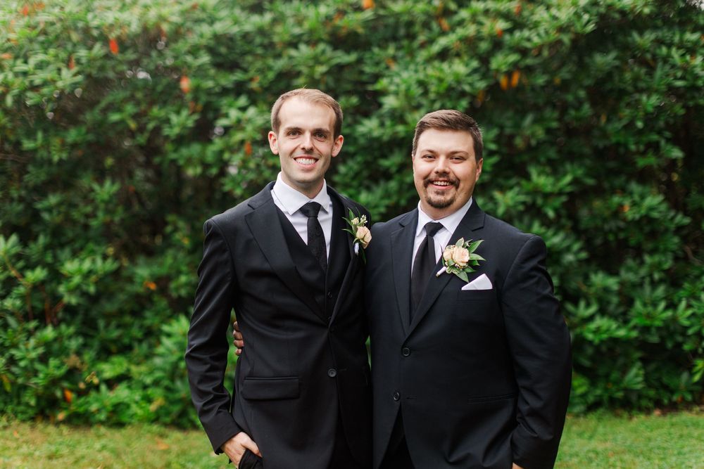 Two groomsmen in black suits with boutonnieres smiling outdoors at a wedding ceremony.