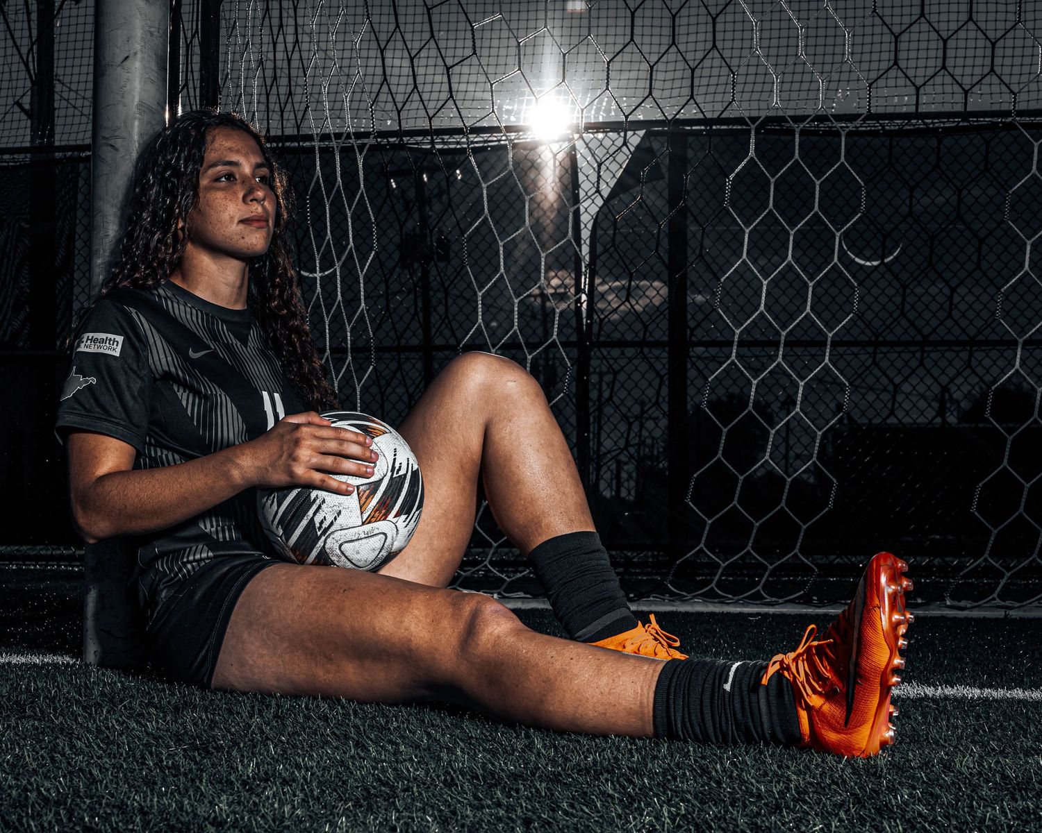 Female soccer player in black jersey sits against chain-link fence holding ball, wearing orange cleats under dramatic lighting.