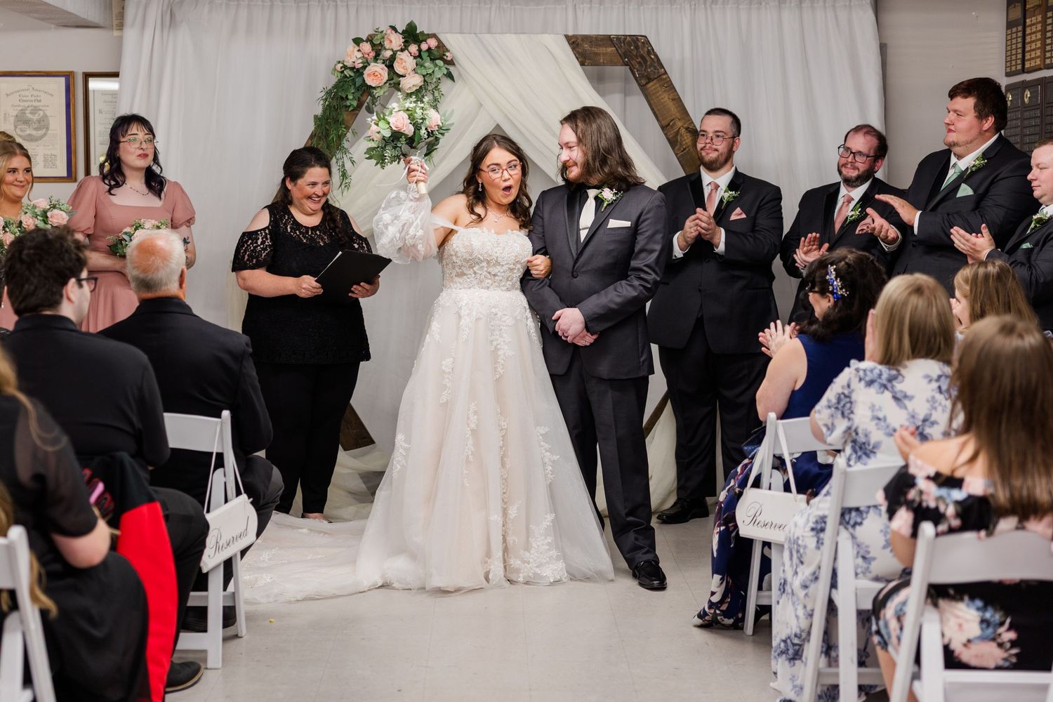 Joyful newlyweds walk down the aisle after wedding ceremony as guests applaud in elegant white venue.