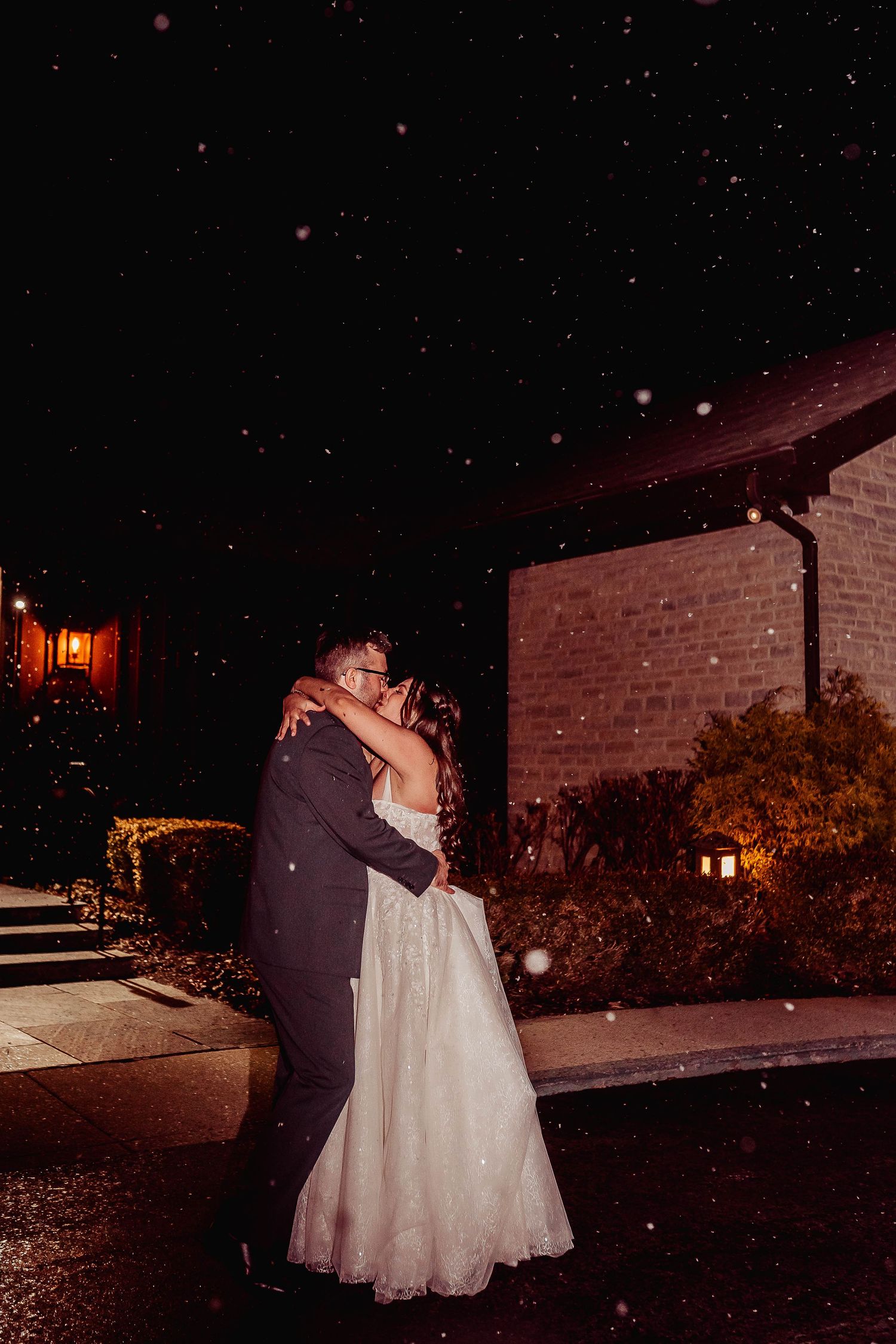 Bride and groom sharing a romantic kiss outdoors at night in the snow, with warm golden lights illuminating the venue.