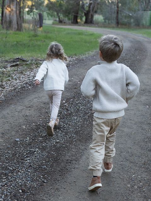 Two young children in cream knit sweaters and tan pants run side by side down a gravel path through a grassy park.