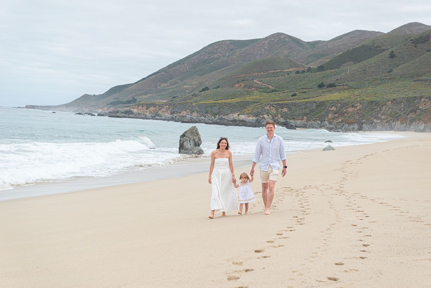 Family of three walking hand-in-hand on a scenic beach with green hills and ocean waves in the background.