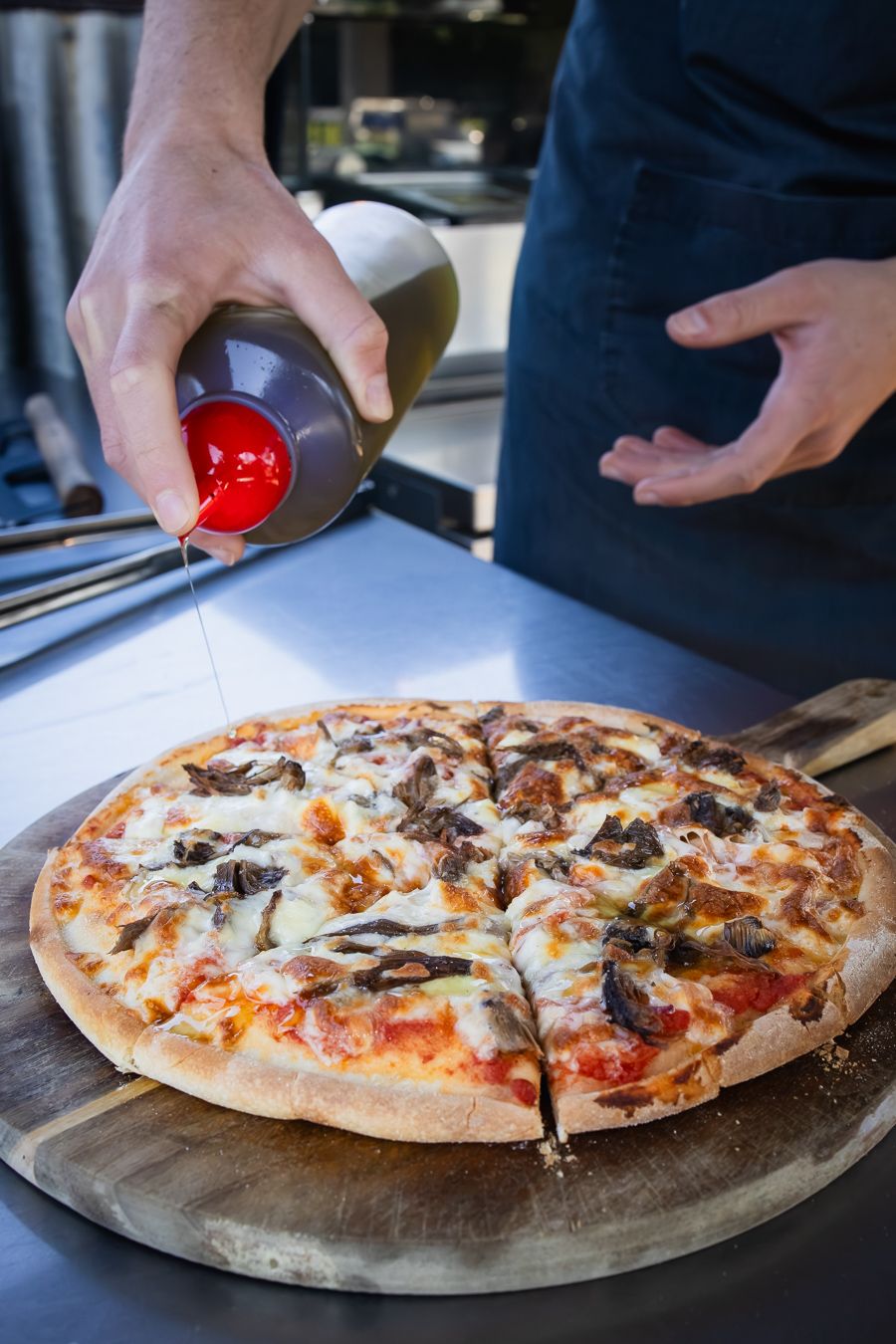 Chef drizzling red sauce over a freshly baked mushroom pizza on a wooden board in a restaurant kitchen.