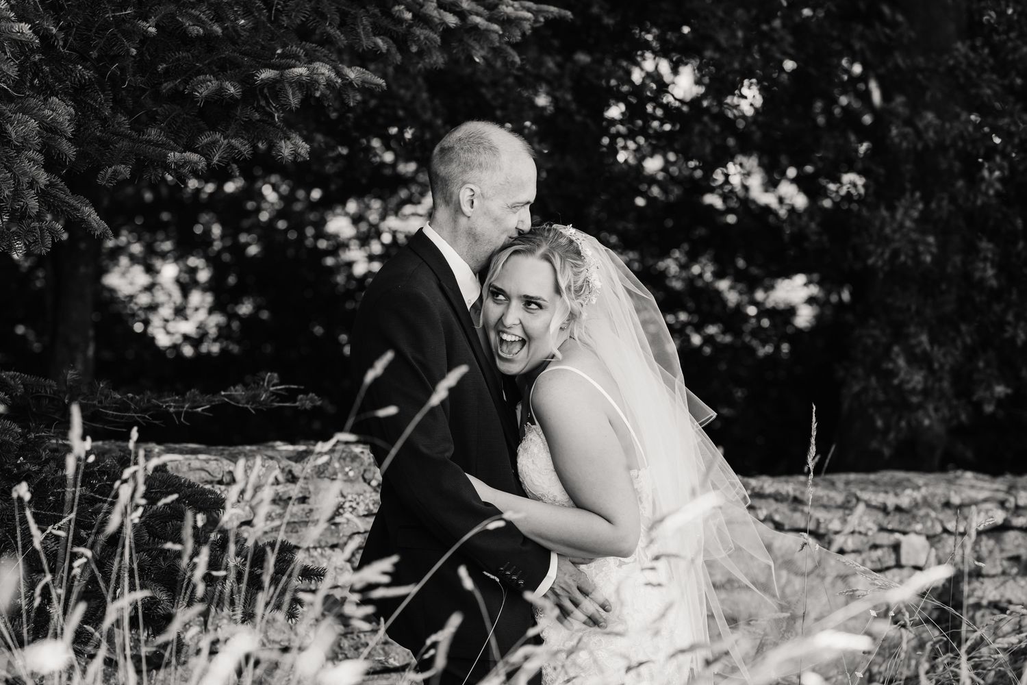 Black and white photo of a smiling bride in veil embracing a groom outdoors among tall grass and trees.