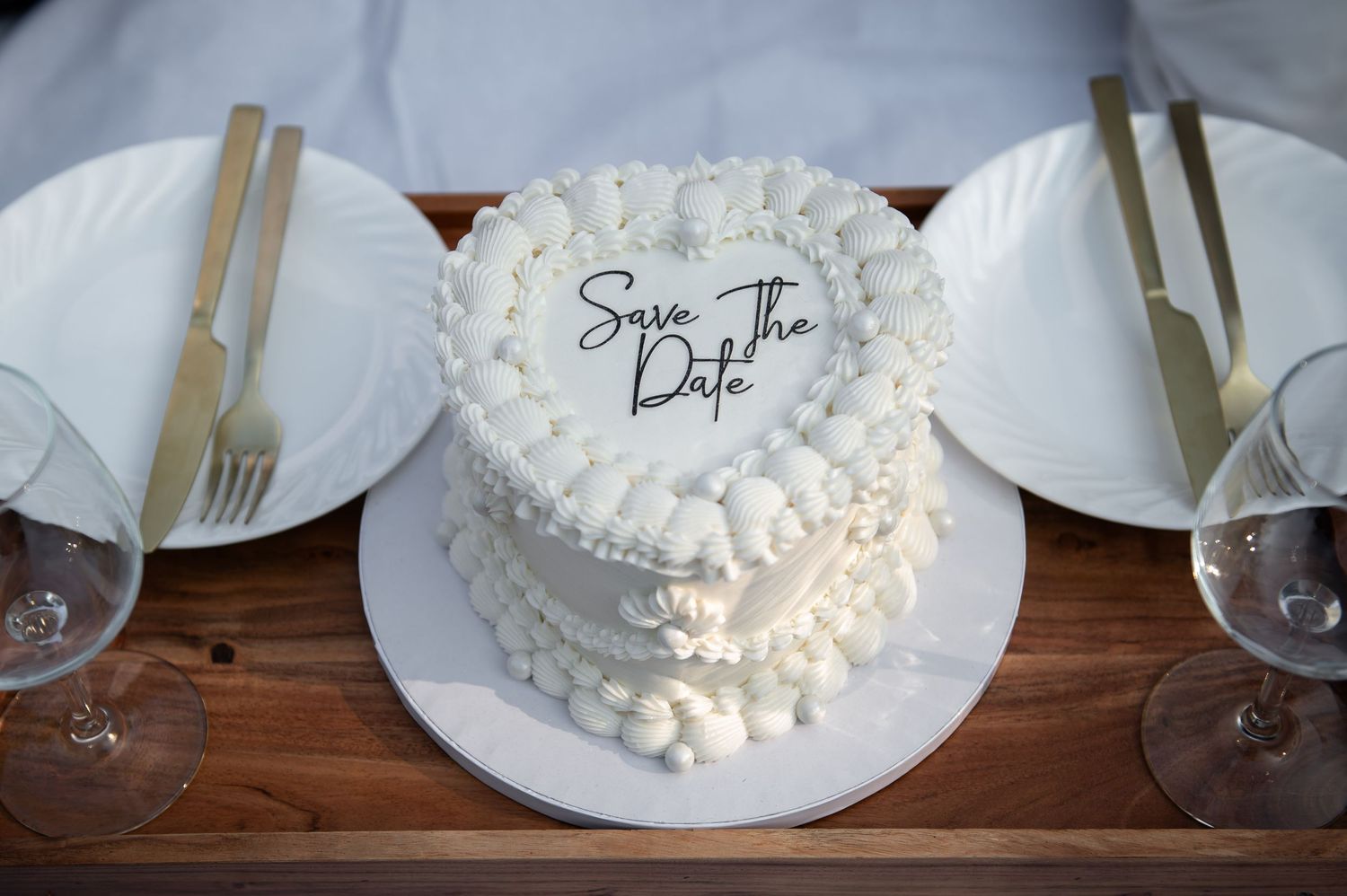 White heart-shaped Save the Date wedding cake with pearl decorations on a wooden table with gold cutlery.