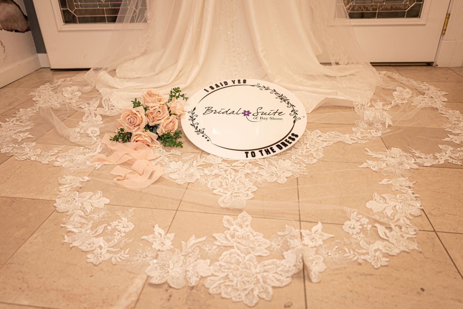 Ivory strapless wedding gown with a long lace-trimmed train hangs on a glass door, with a peach rose bouquet resting on the tile floor.