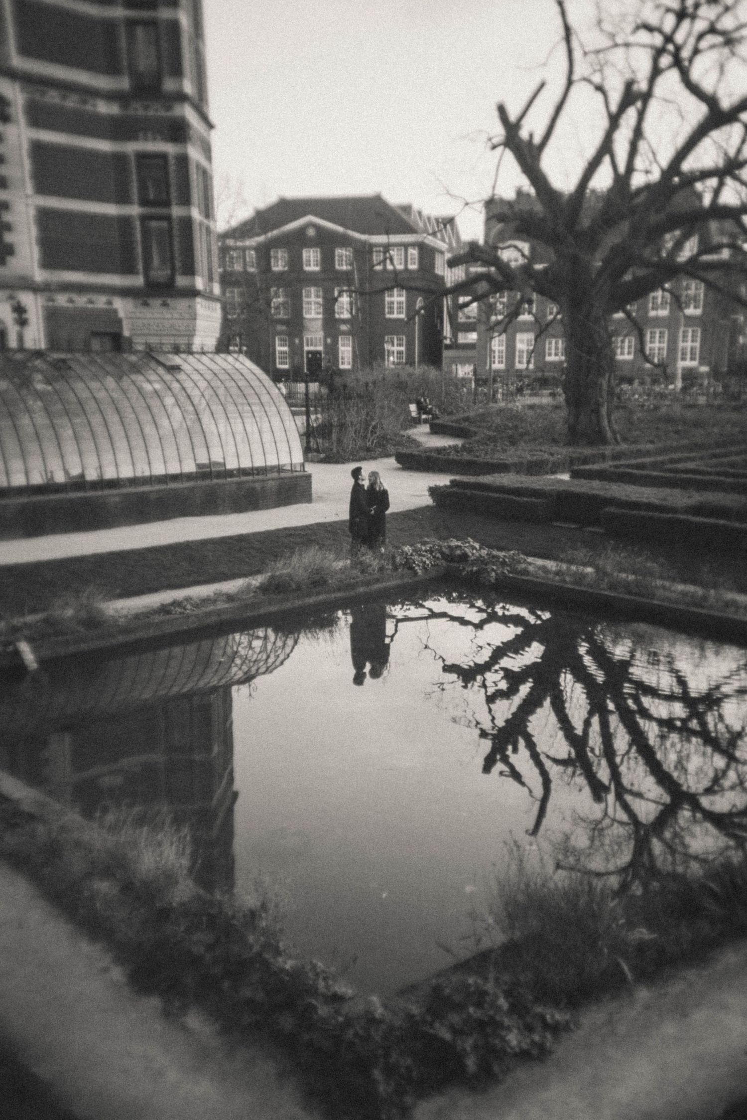Black and white photo of two people standing beside a reflective pond near a greenhouse, Rijksmuseum Amsterdam.