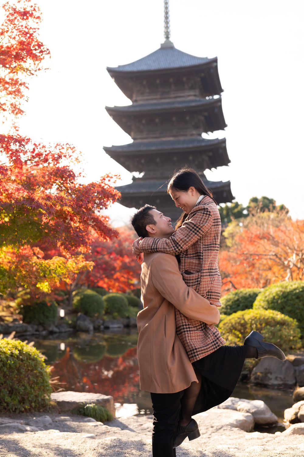 Couple embracing in a Japanese garden as he lifts her, with a five-story pagoda, pond, and red autumn maple leaves behind them.