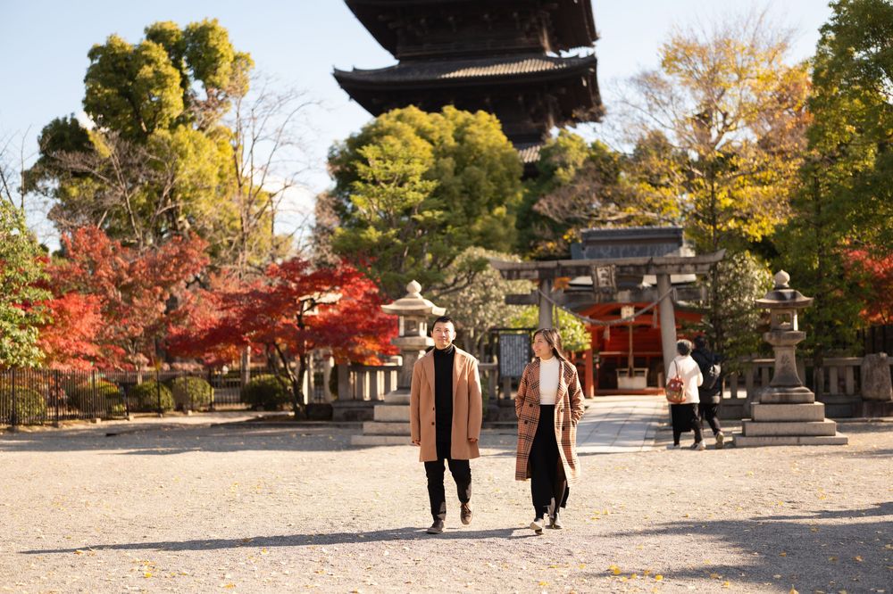 Two people in long coats walk across a gravel courtyard at a Japanese temple with a pagoda and vivid autumn foliage.