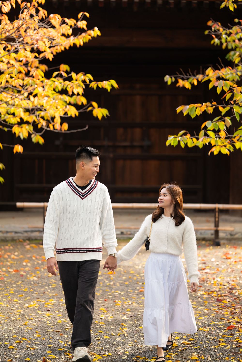 Smiling couple holding hands and strolling beneath golden autumn leaves in front of a traditional wooden building.