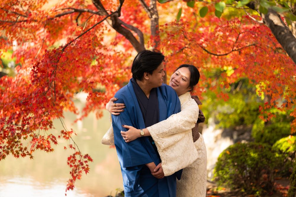 Couple in traditional Japanese kimono embracing under vibrant red autumn maple leaves beside a calm lake.