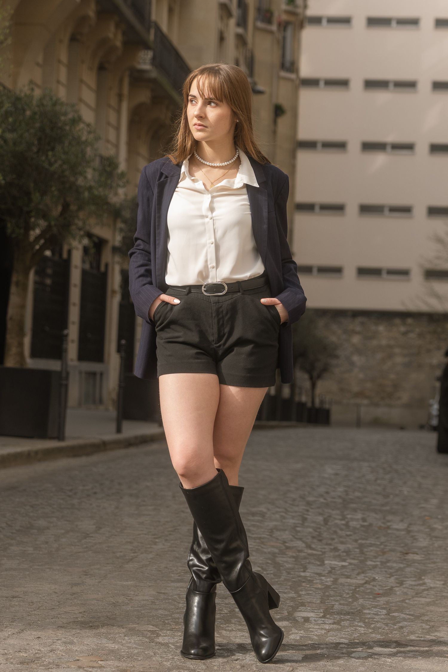 Portrait lifestyle d’une femme en blazer marine et bottes noires dans une rue pavée de Paris