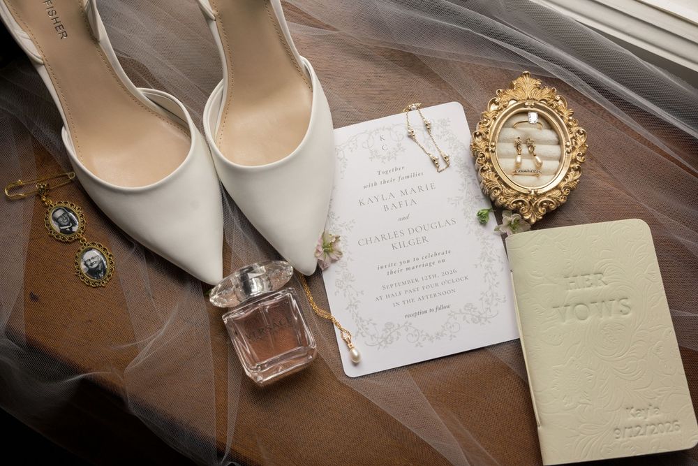 Elegant wedding flat lay featuring white heels, perfume, gold jewelry, floral invitations, and a vow booklet on sheer fabric.