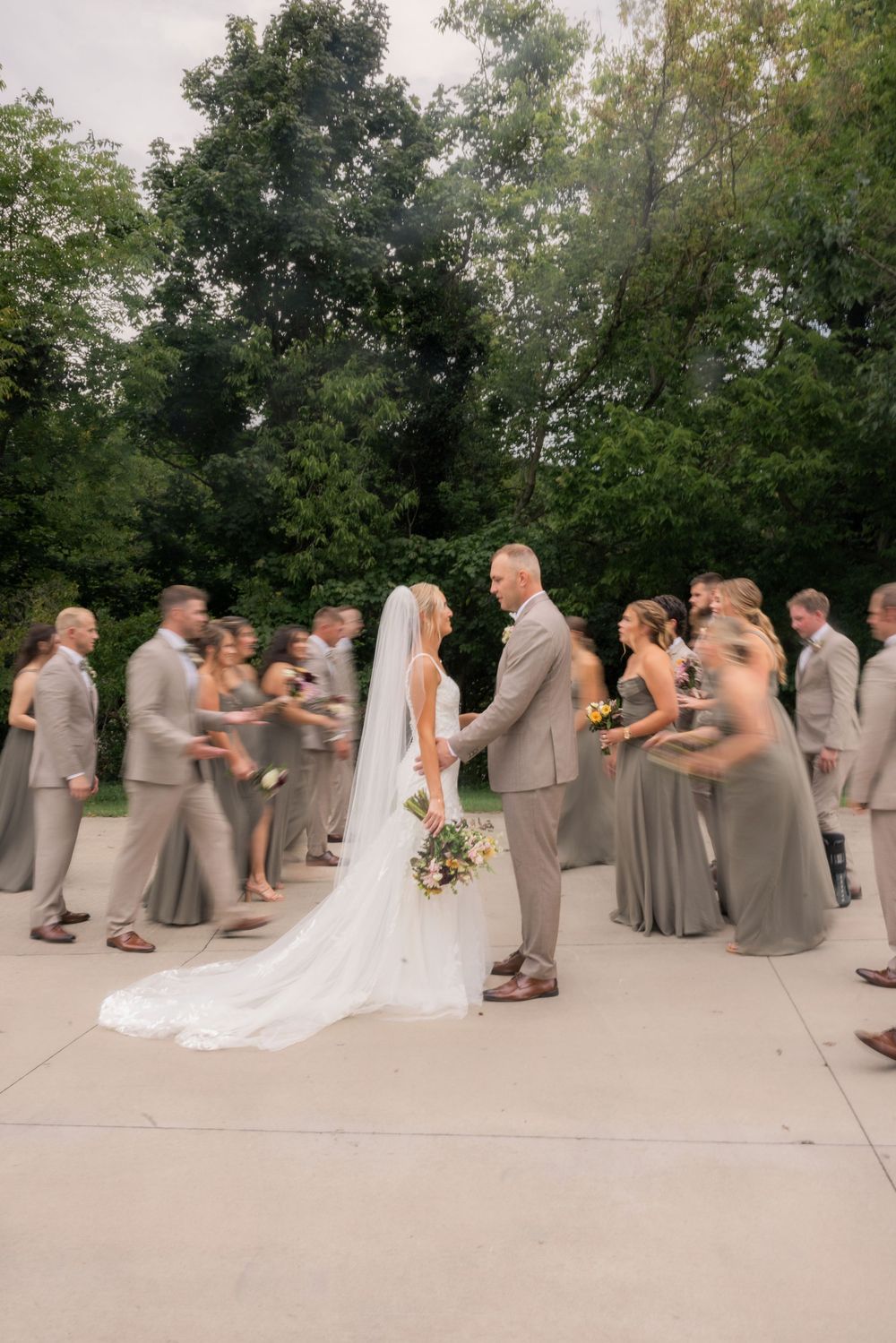 Bride and groom hold hands surrounded by wedding party in sage green attire on a paved outdoor venue with lush trees.