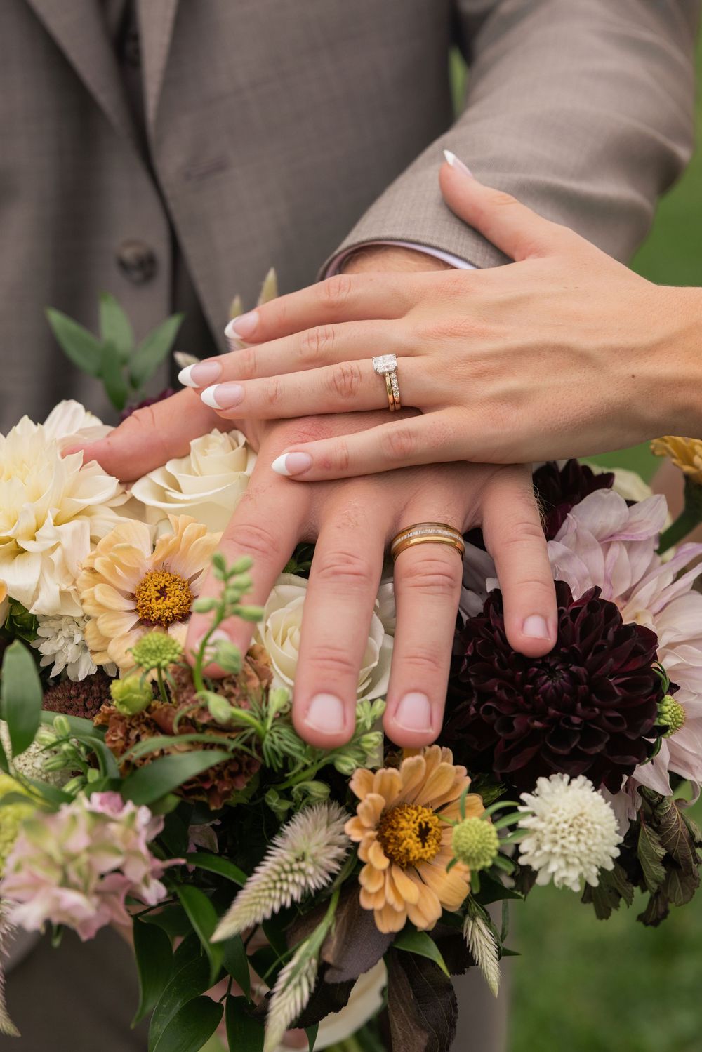 Newlyweds display wedding rings on hands resting over a colorful bridal bouquet with dahlias and roses.