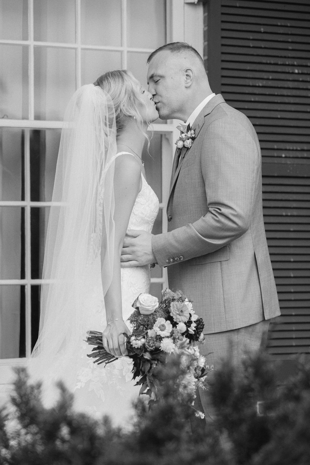 Black and white photo of a bride and groom sharing a kiss, bride holding a bouquet, standing outside a building.