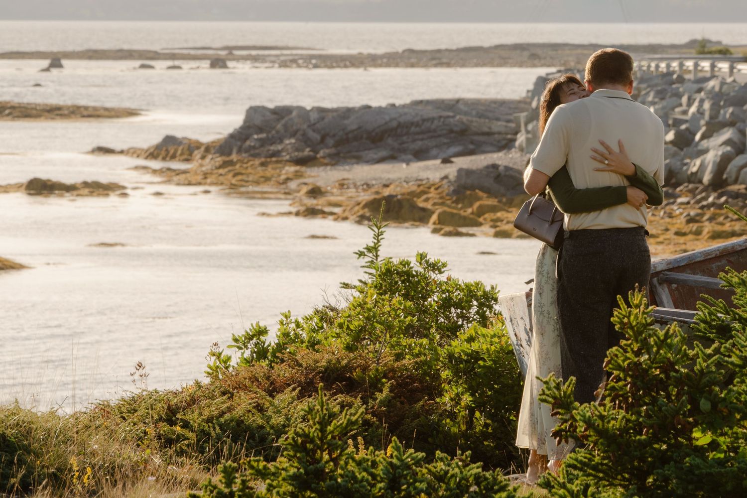 Couple embracing while overlooking a scenic rocky coastline with calm ocean waters and lush greenery.
