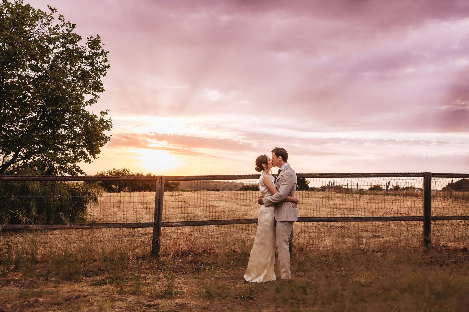 Couple sharing a romantic kiss at sunset by a rustic wooden fence in a golden field.