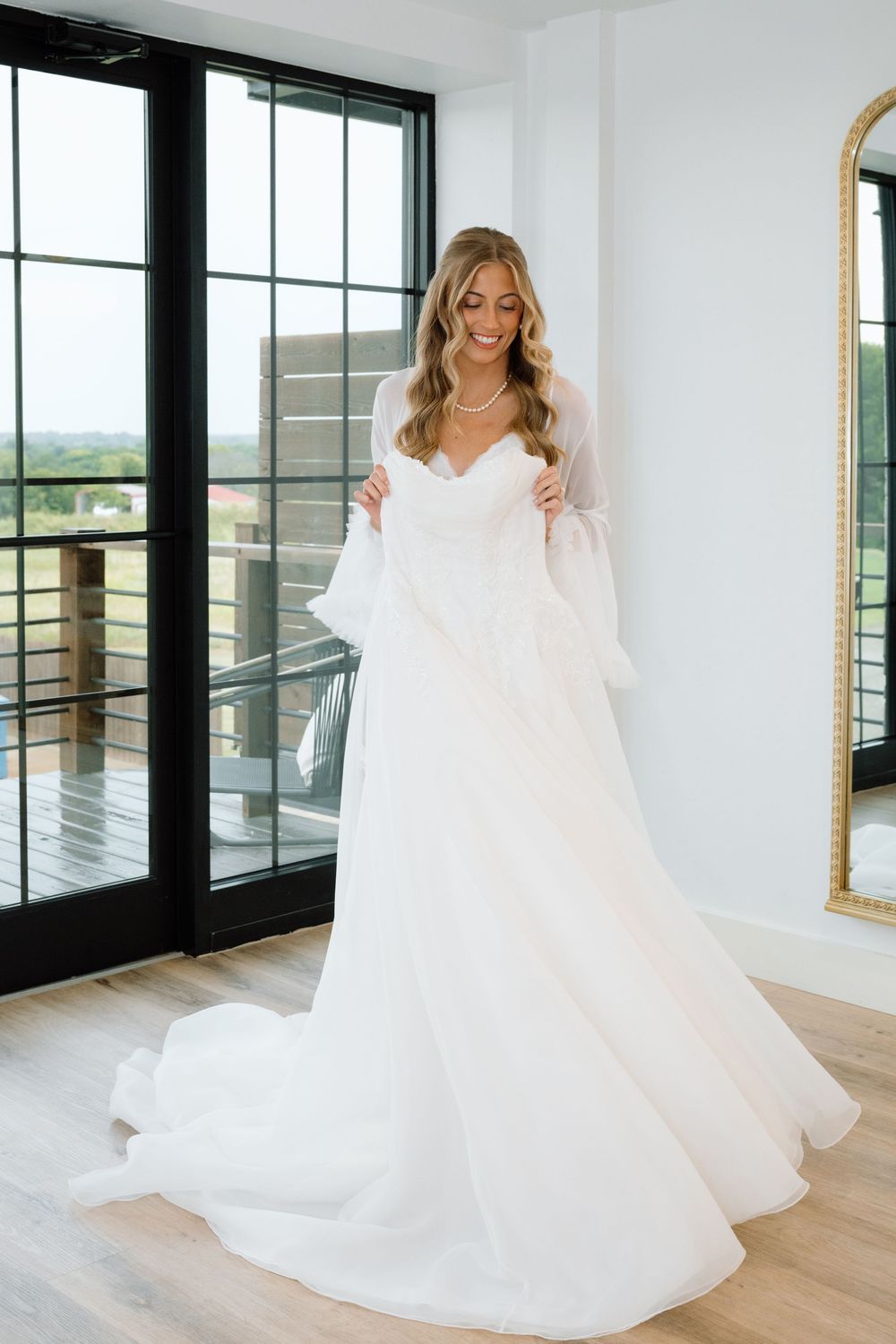 Bride in a flowing white A-line wedding gown with bell sleeves stands near French doors, smiling softly in a bright room.