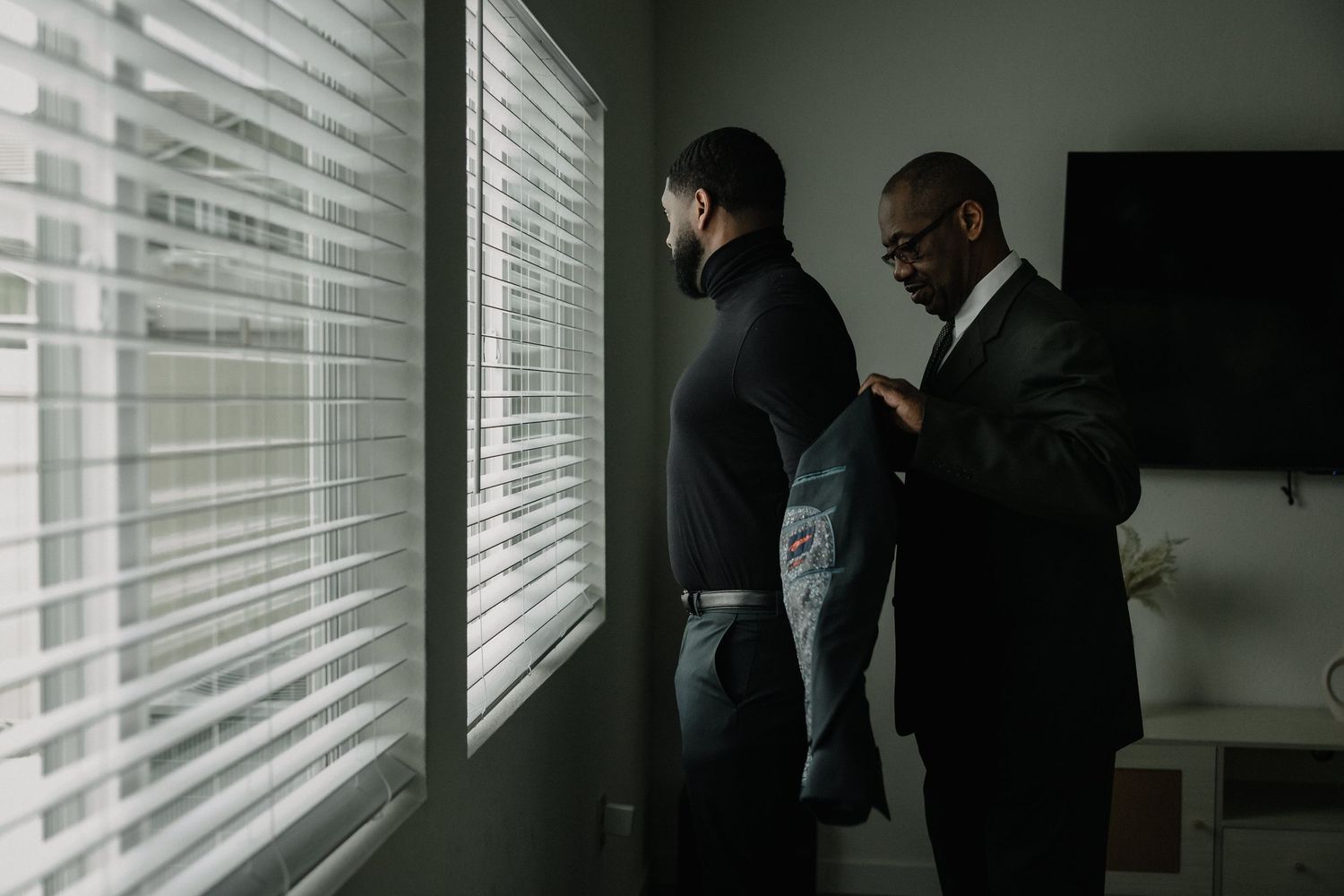 Two men in formal black suits stand by window blinds, one helping the other adjust his jacket in a well-lit room.