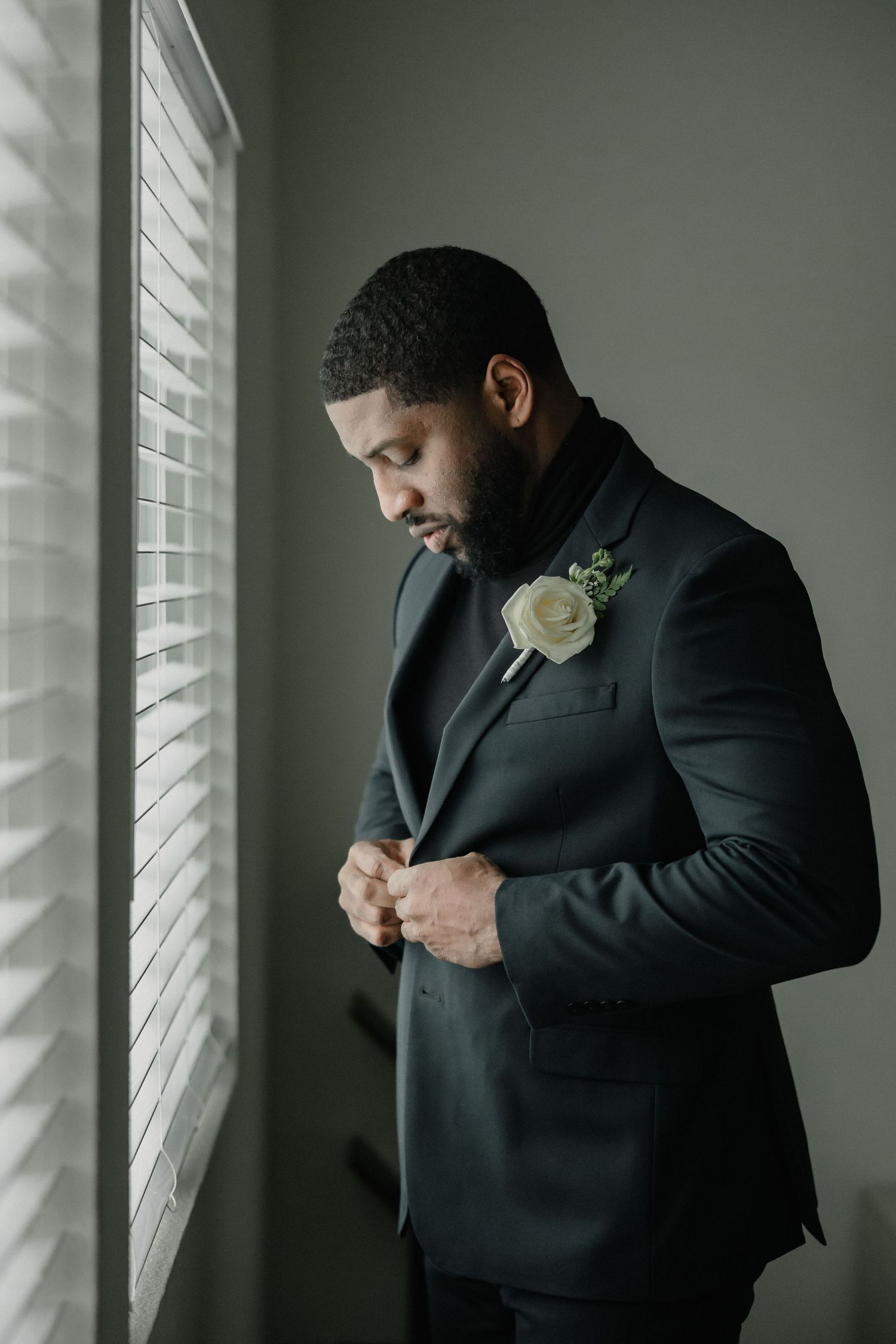 A groom in a dark suit with a white rose boutonniere buttons his jacket while standing by a window.