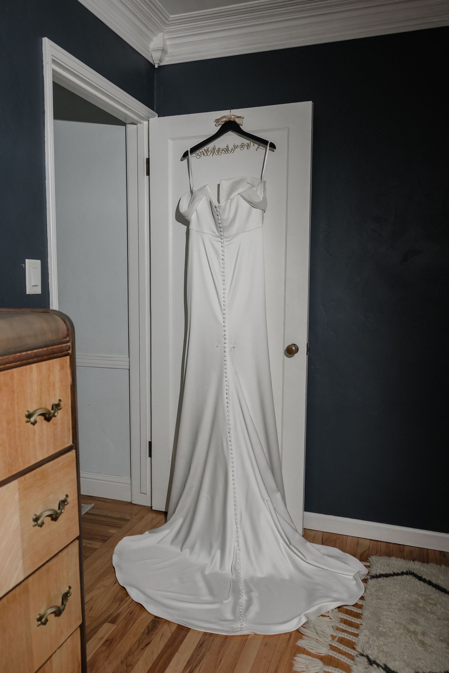 Elegant white satin wedding gown with button back detail hanging on a door in a navy blue bedroom.