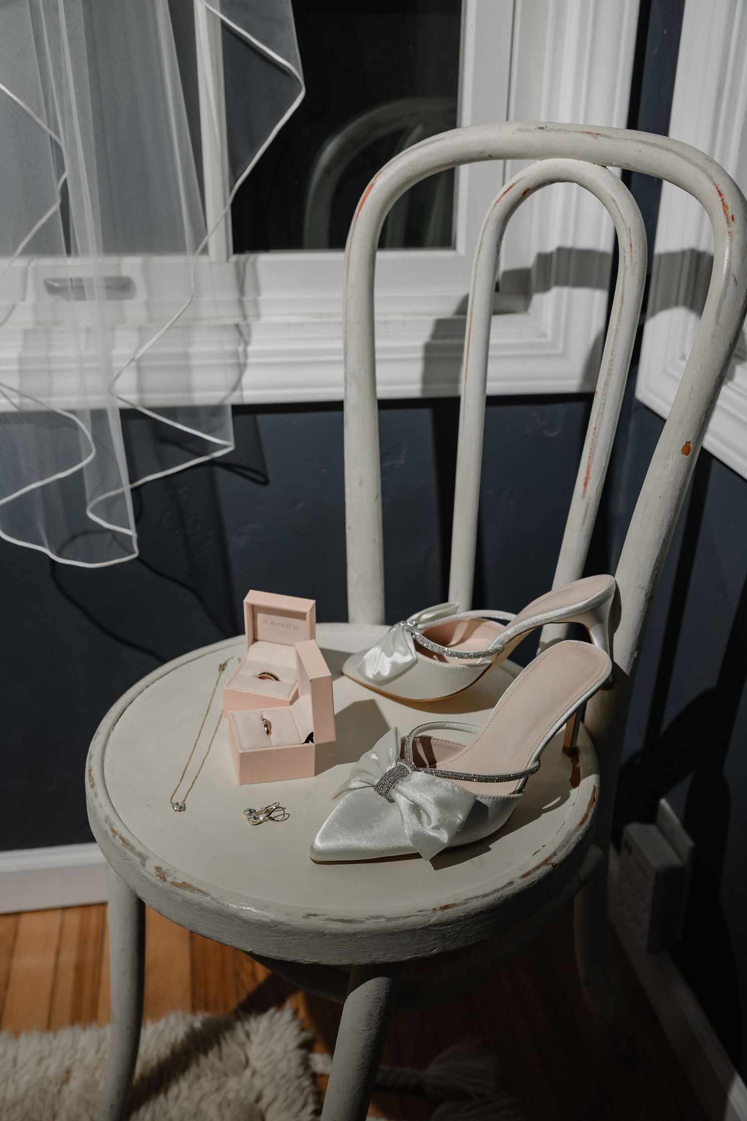 Elegant bridal heels with satin bow and jewelry boxes displayed on a vintage white wooden chair near a window.