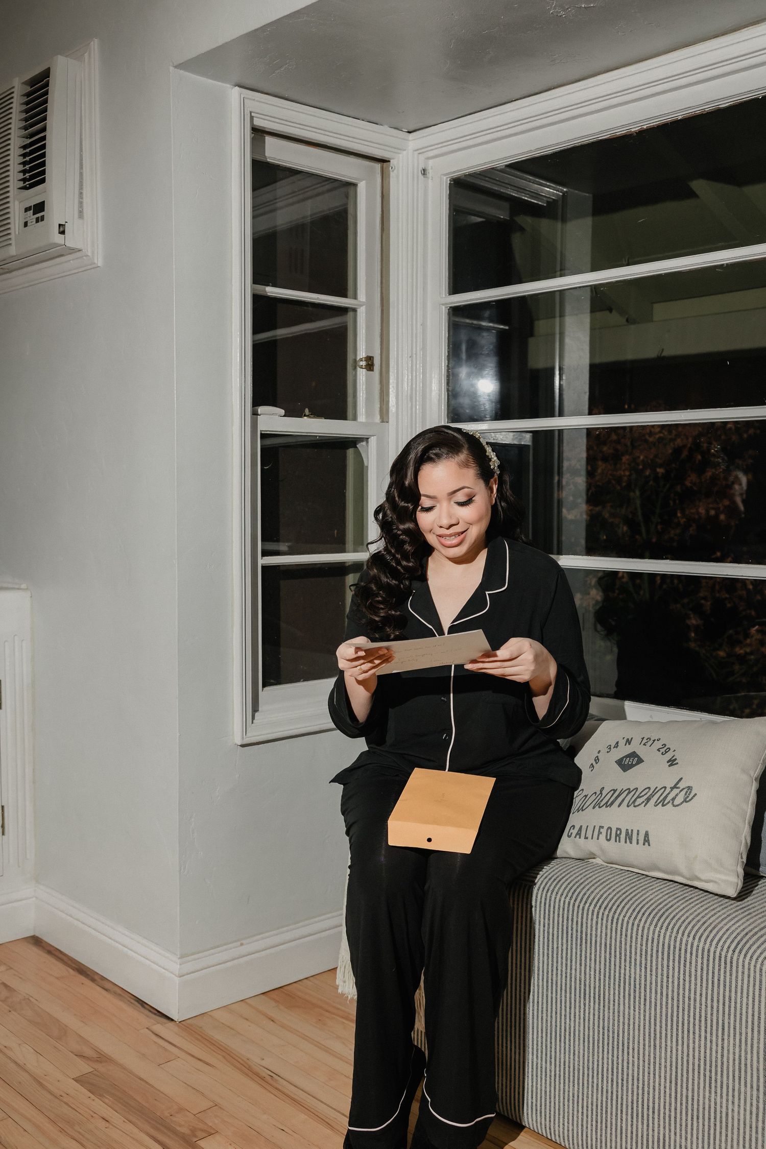 Woman in black pajamas reading a card while holding a wooden gift box near a window at night.