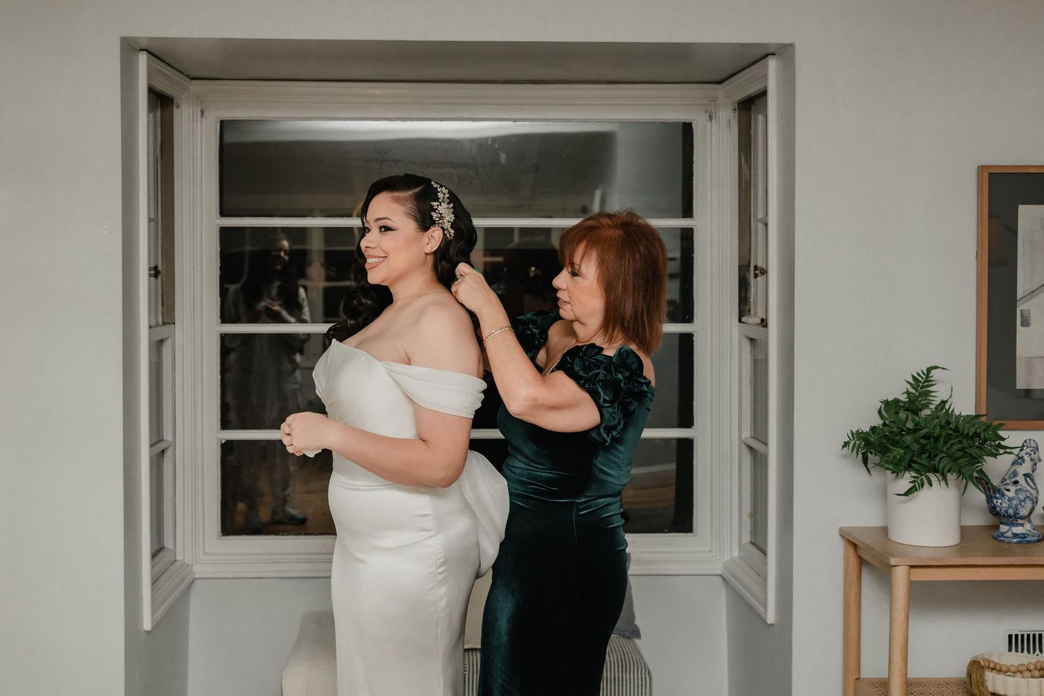 Mother helping bride button up white wedding dress near French doors before ceremony.
