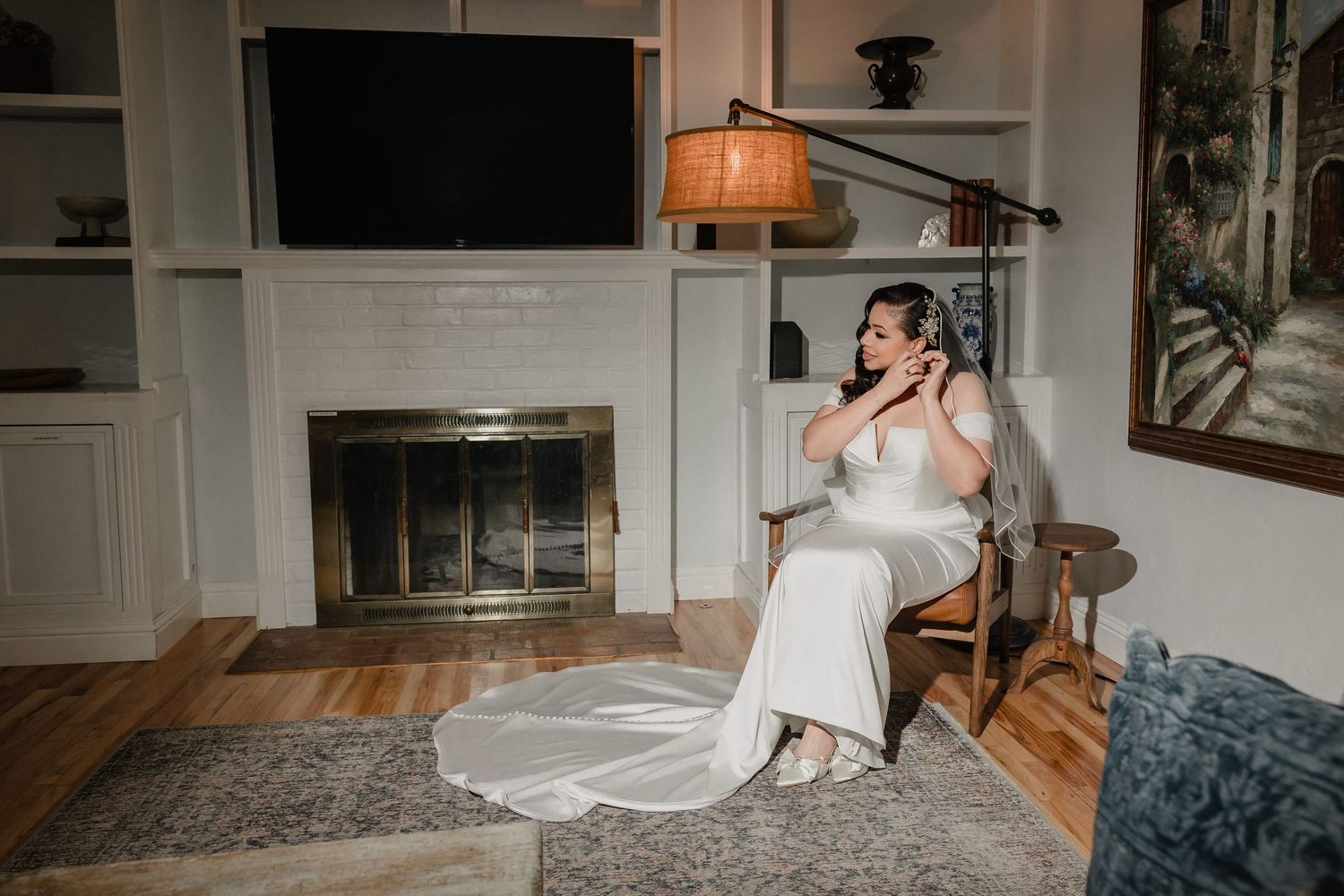 Bride in white wedding gown sits by fireplace in elegant room, adjusting earrings before ceremony.