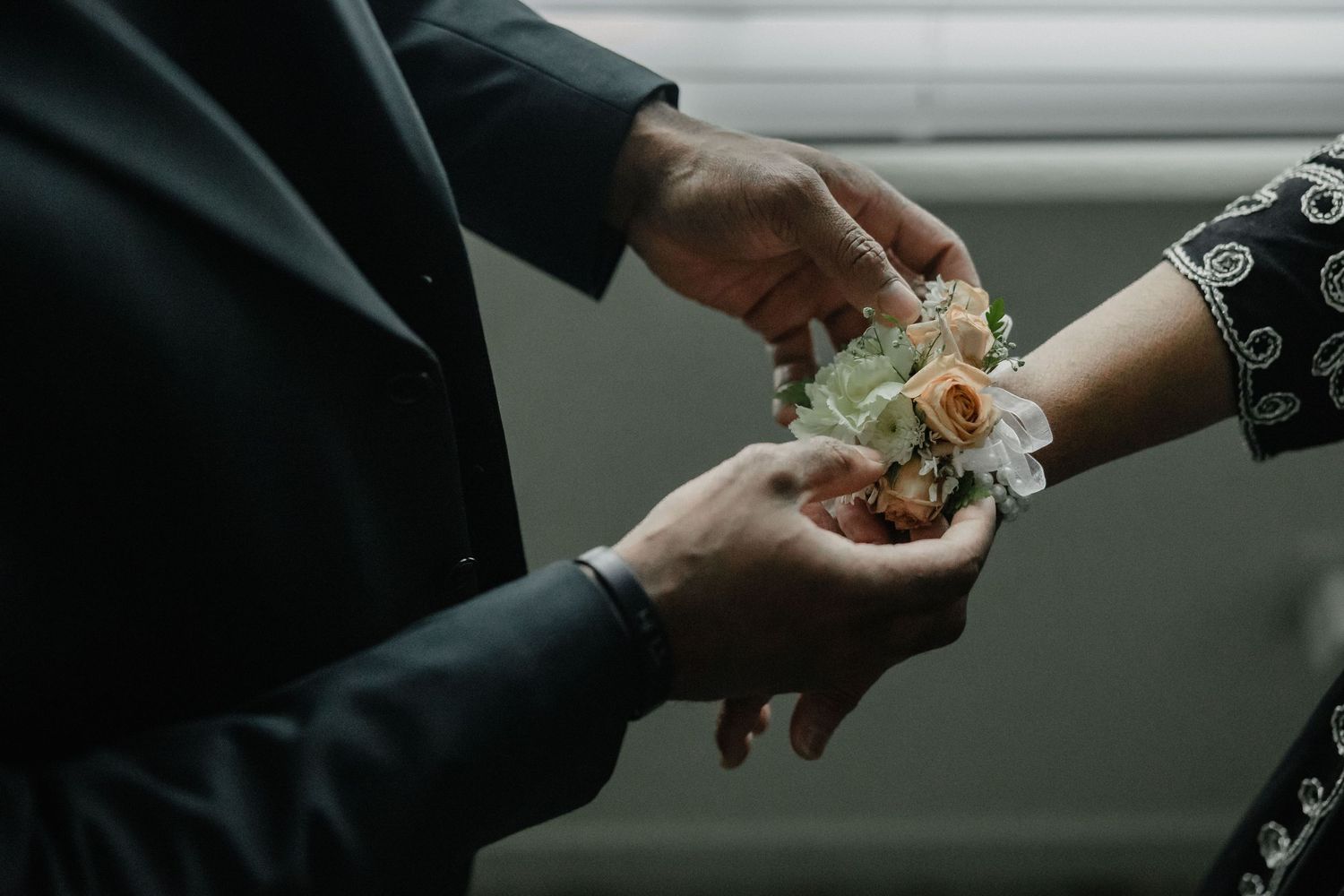 A groom carefully placing a floral wrist corsage with peach roses on a bride's wrist at a wedding ceremony.