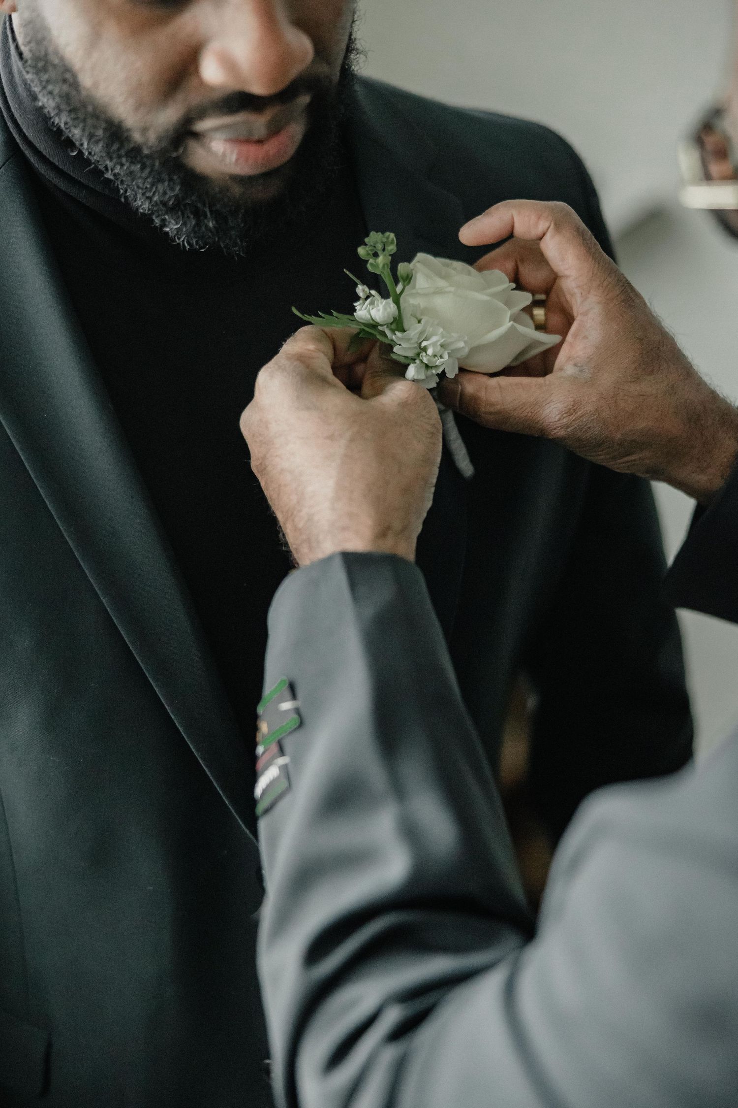 A groomsman pins a white rose boutonniere onto a man in a black suit jacket at a wedding ceremony.