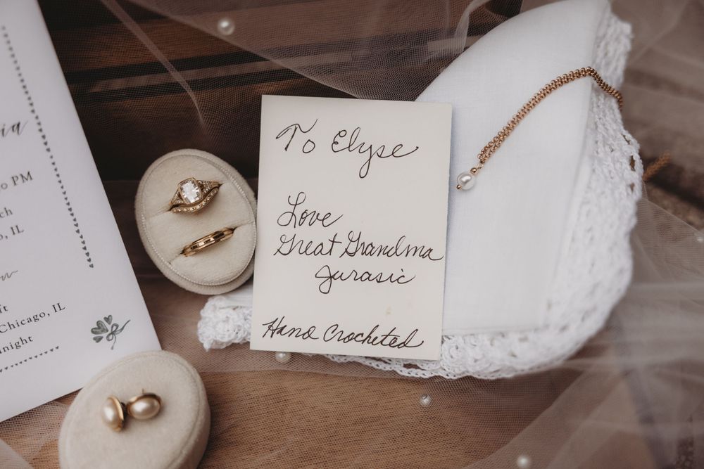 Vintage wedding flat lay with gold rings, handwritten note from great grandma, and hand-crocheted lace accessory on wooden surface.