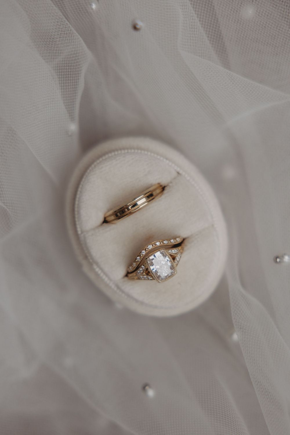 Gold wedding ring set with diamond engagement ring displayed in ivory velvet box on white bridal veil with pearls.