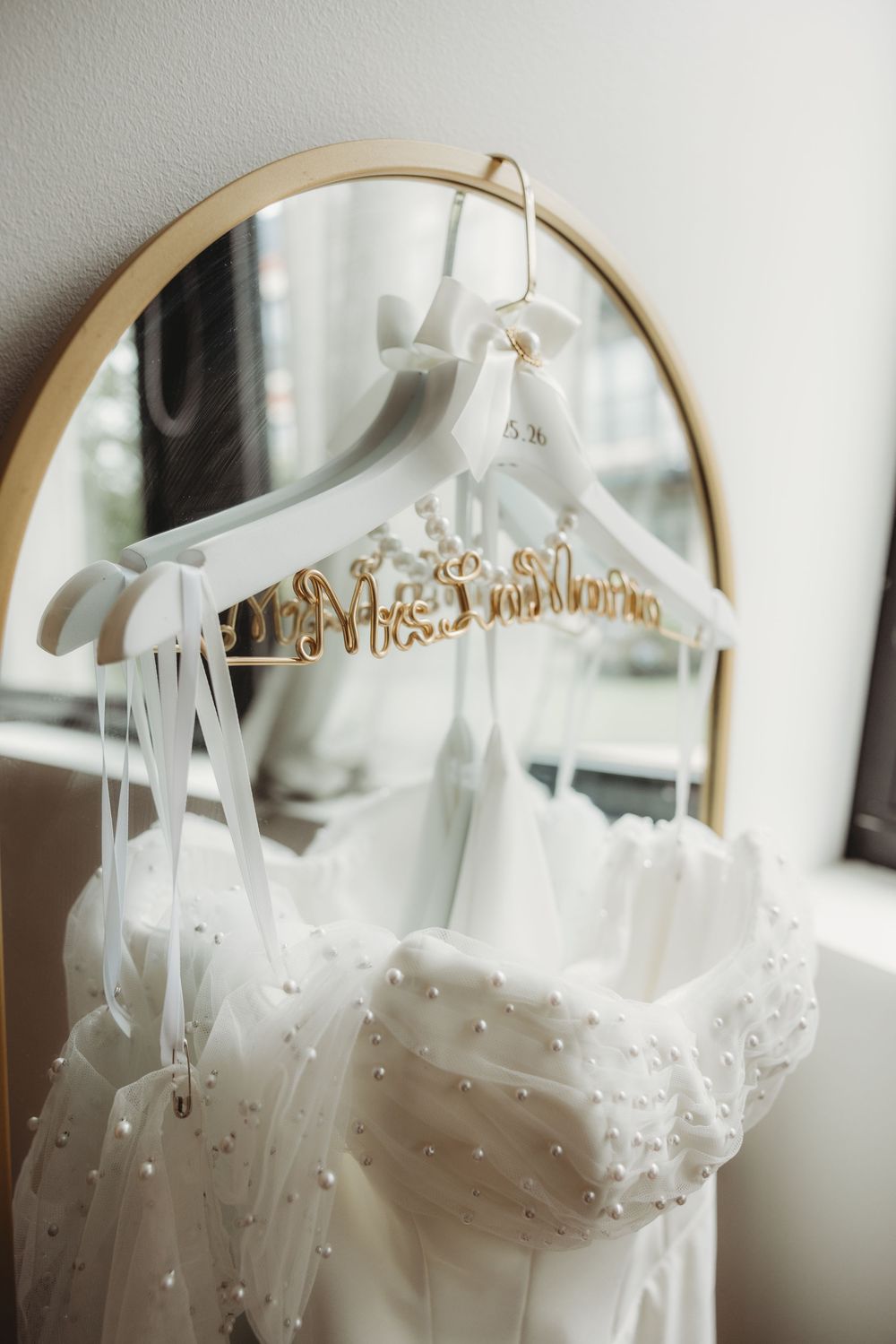 White pearl-embellished wedding dress on custom Mrs. hanger in front of gold arch mirror.