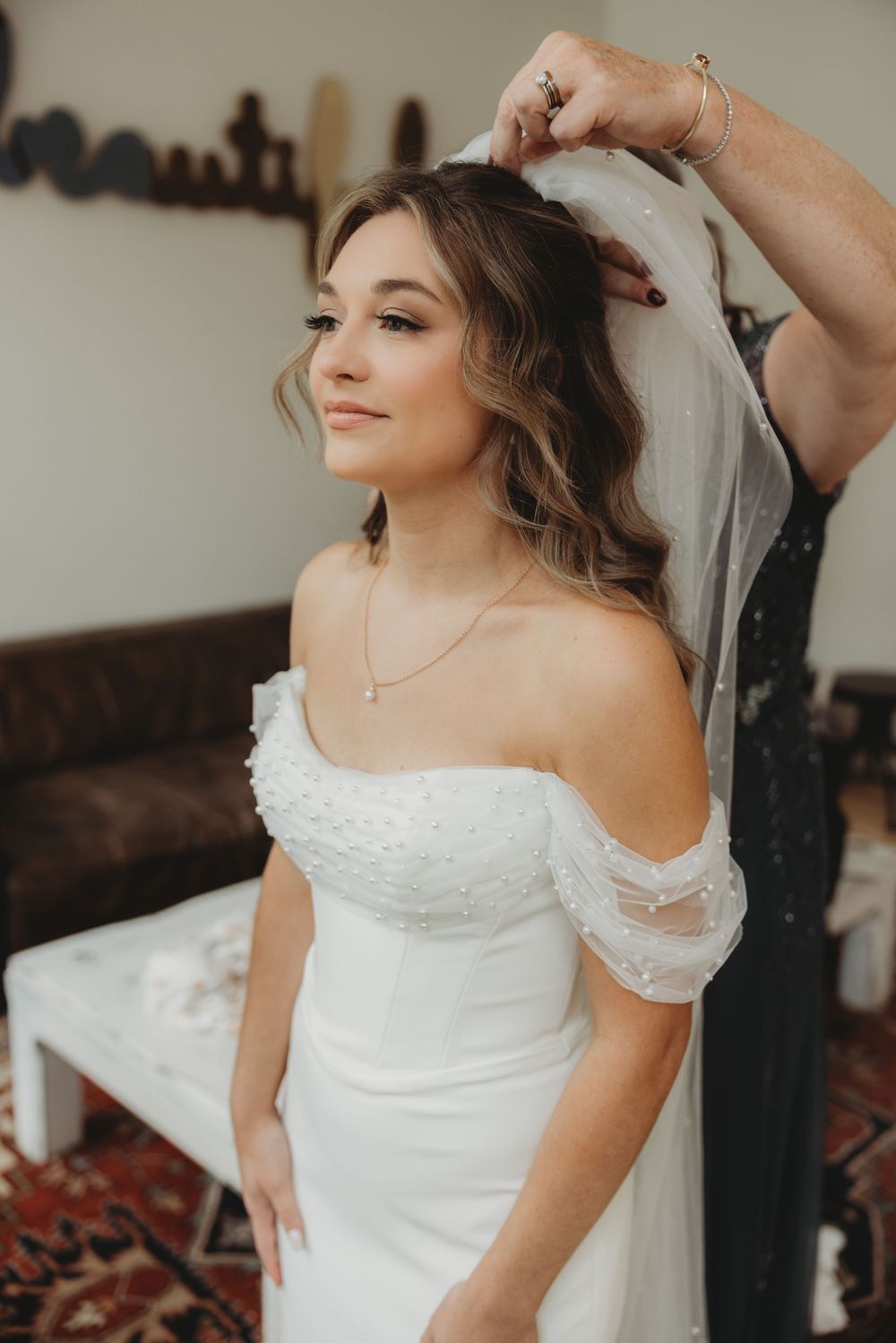 Bride in pearl off-shoulder wedding gown getting veil placed by assistant.