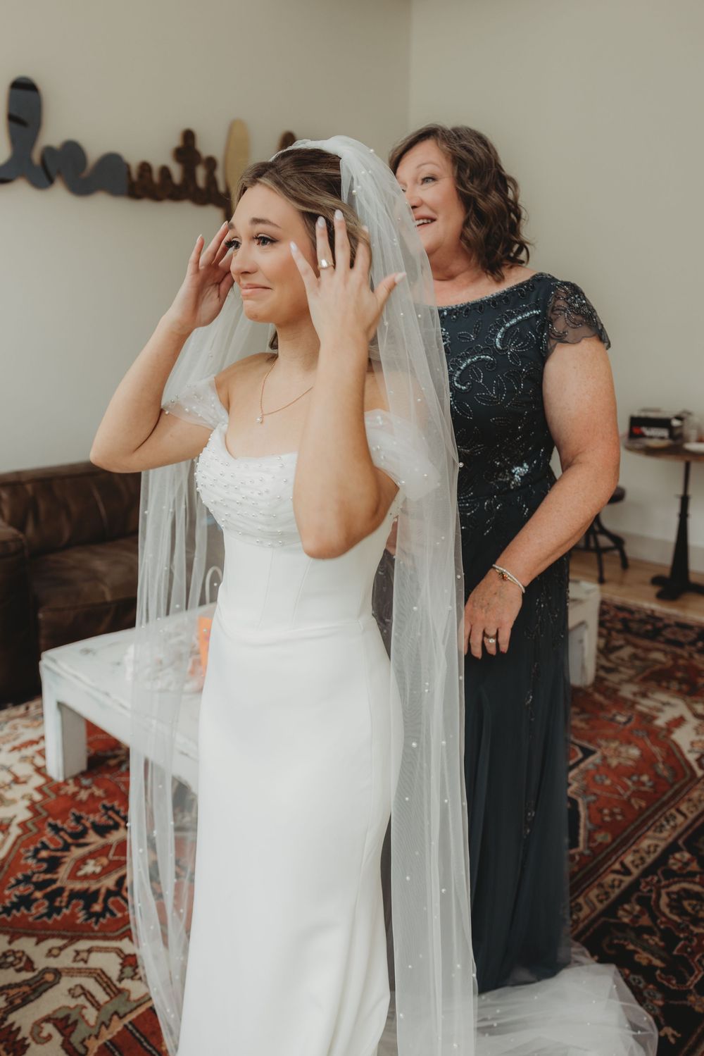 Bride in white gown adjusts her veil while a woman in a dark dress assists her during wedding preparations indoors.