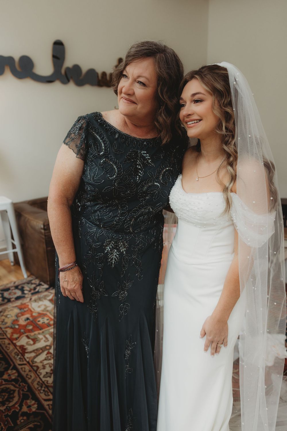 A bride in a white gown and veil smiles beside an older woman in a dark beaded gown at a wedding.
